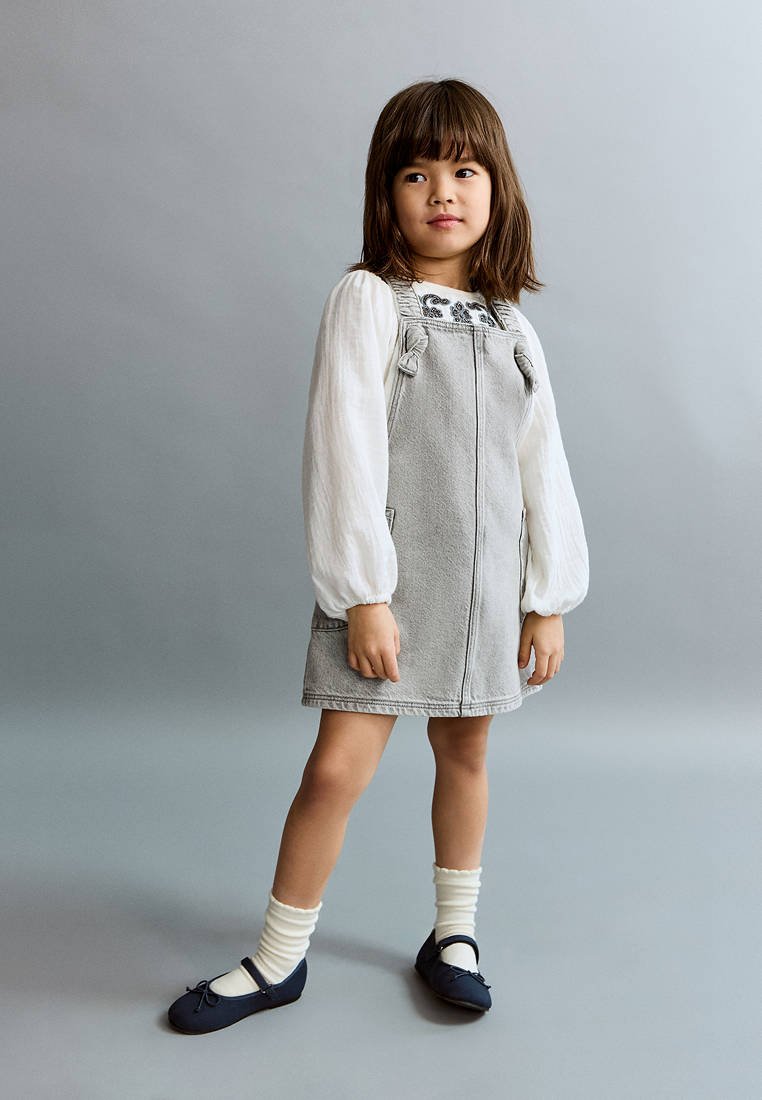 Jeune fille aux cheveux bruns portant une robe tablier grise sur un chemisier blanc, des chaussettes blanches et des chaussures bleu foncé, debout sur un fond gris uni.