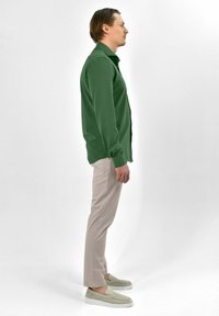 Groene lange mouwen shirt, lichtbeige broek en grijze instappers. Het shirt heeft een knoopsluiting en een ontspannen pasvorm, wat een casual stijl toont in zijaanzicht.