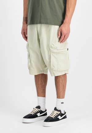 Mann trägt hellbeige Cargoshorts, schwarz-weiße Sneakers, weiße Socken und ein olivgrünes Hemd, steht vor einem weißen Hintergrund.