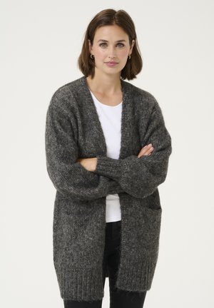 Cardigan en tricot gris avec des manches larges, ouverture à l'avant et deux poches avant. Porté sur un haut blanc, il présente une texture douce et une coupe décontractée.
