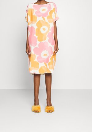 Frau trägt ein locker sitzendes, knielanges Kleid mit großen pinken und orangen Blumenmustern und gelben, flauschigen Pantoletten mit offenen Zehen, steht vor einer schlichten Wand.