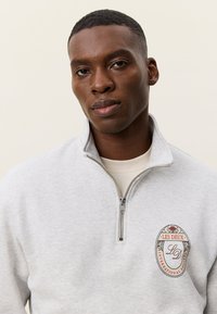 Γκρι φούτερ με φερμουάρ και ψηλό γιακά, με διακοσμητικό σήμα σε οβάλ σχήμα με λουλουδένιες λεπτομέρειες και κείμενο. Κατασκευασμένο από μαλακό ύφασμα.