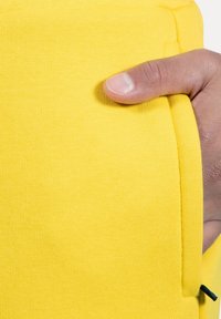 Żółty materiał tekstylny o gładkiej fakturze. Ręka trzyma boczną kieszeń; kieszeń ma kontrastujący ciemny akcent wzdłuż krawędzi.