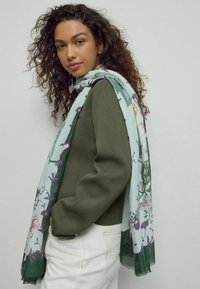 Zielony sweter wykonany na drutach w połączeniu z jasnoniebieskim szalikiem w kwiatowy wzór z fioletowymi i zielonymi akcentami. Szalik jest luźno zarzucony.