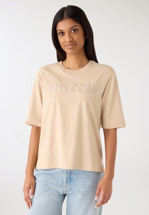 Giovane donna con lunghi capelli scuri che indossa una maglietta beige con il testo "CAPPUCCINO" e jeans azzurri su sfondo bianco.