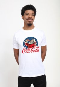 Hvid bomulds t-shirt med et vintage Coca-Cola grafik af julemanden i rødt tøj. Designet inkluderer stjerner og en blå cirkulær baggrund.