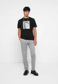Zwart T-shirt met een grafische print van een tijger in wit, gecombineerd met lichtgrijze broeken en zwarte loafers. Accentuering met een zilveren ketting.