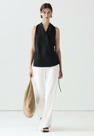 Donna che cammina con top nero senza maniche a incrocio, pantaloni bianchi ampi, sandali neri, che tiene una grande borsa morbida marrone chiaro su sfondo neutro.