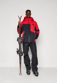 Skioutfit mit einer rot-schwarzen wasserdichten Jacke mit Reißverschluss und passenden schwarzen Hosen. Hält Skier und trägt schwarze Skischuhe.