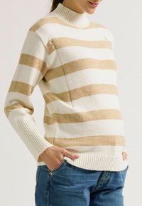 Gestreifter Strickpullover in Creme und Beige mit hohem Kragen, gerippten Bündchen und lockerer Passform, kombiniert mit blauen Jeans.