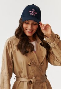 Căciulă de baseball navy cu text brodat roz "MY F❤️RVER DATE", purtată de o femeie cu păr lung, asortată cu o trench coat bej.