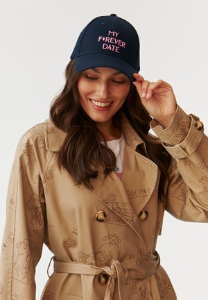 Navy baseball cap with pink embroidered text "MY F❤️RVER DATE," worn by a woman with long hair, paired with a beige trench coat.