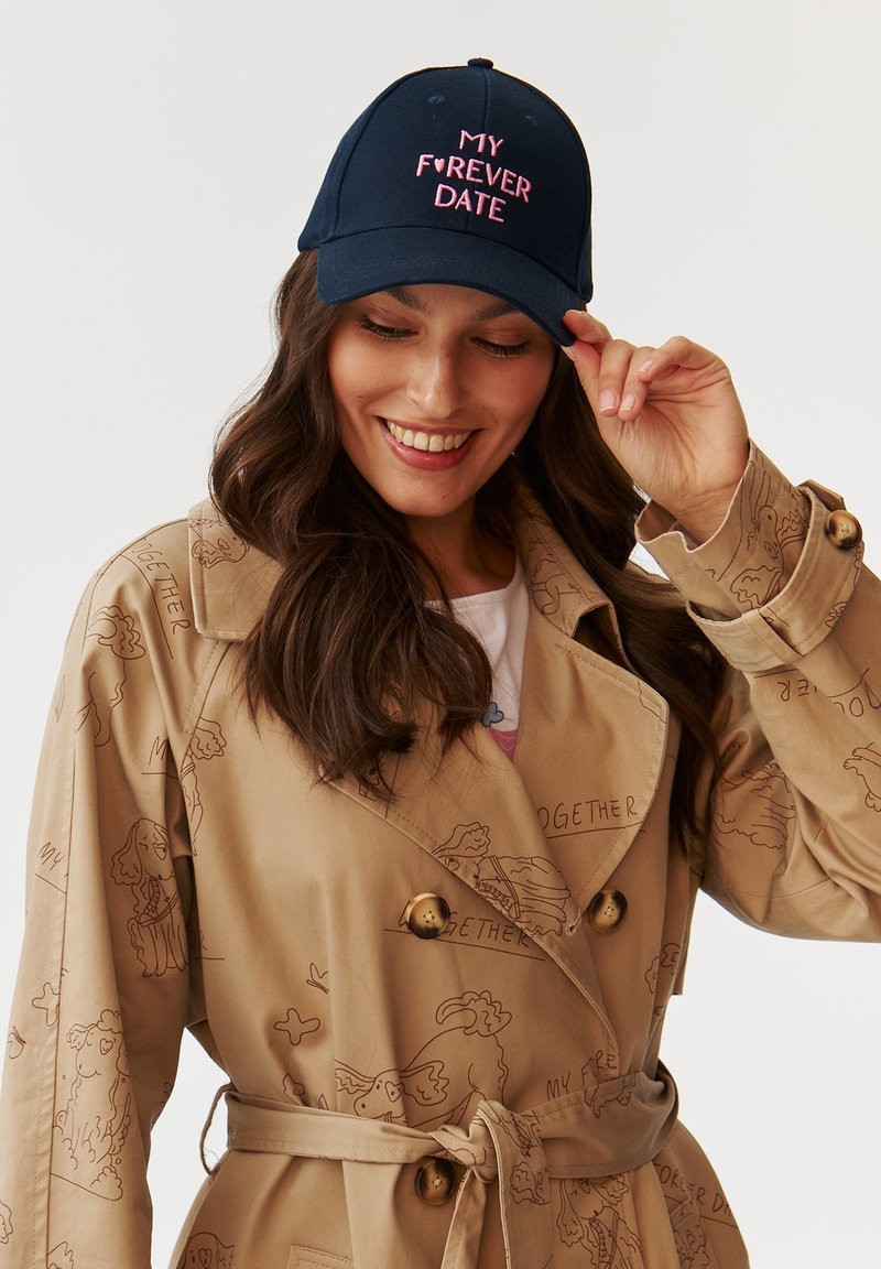 Căciulă de baseball navy cu text brodat roz "MY F❤️RVER DATE", purtată de o femeie cu păr lung, asortată cu o trench coat bej.