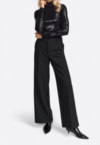 Donna che indossa un top lungo a maniche nere lucide con paillettes, pantaloni neri a vita alta e larghi, e stivali neri con punta a punta e tacco.