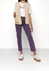 Beige fleecepaita, jossa painonapit, valkoisen T-paidan päällä, yhdistetty violetteihin farkkuihin ja kermaan sävyisiin lenkkareihin, joissa on ruskea pohja.