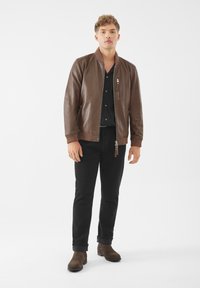 Giacca bomber in pelle marrone con polsini e collo a costine, zip frontale e tasca decorativa con zip. Abbinata a pantaloni neri e stivali marroni.