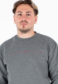 Giovane uomo con capelli corti e ondulati che indossa una felpa grigia con il piccolo testo rosso "BIKKEMBERGS" centrato sul petto, espressione neutra.