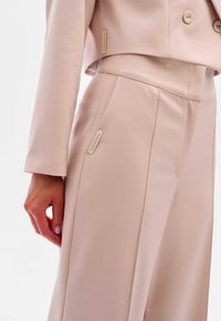 Beige cropped jasje en bijpassende broek van gladde stof. Opvallende details zijn de merklabels en de op maat gemaakte naden.