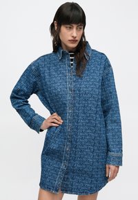 Sininen farkkupaitamekko, jossa toistuva logokuvio, napitettu suljenta ja löysä istuvuus sivuhalkioilla, pitkät hihat ja kaulus.