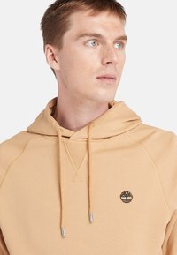Beige hoodie med snoretræk, med et træ-logo på venstre bryst. Lavet af blødt stof med en glat tekstur og raglanærmer.