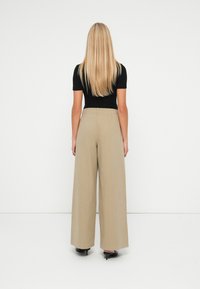 Mulher loira de costas, vestindo uma blusa preta de manga curta, calças largas bege e sapatos de salto preto com bico fino, sobre fundo branco.