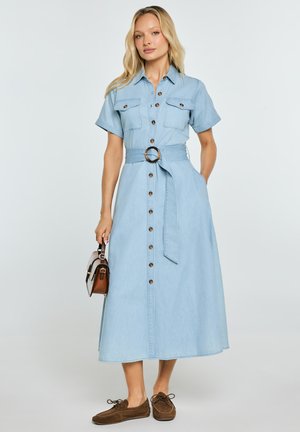 Kobieta ubrana w jasnoniebieską sukienkę midi na krótki rękaw z guzikami z przodu, z kieszeniami i paskiem w talii, trzymająca małą brązową torebkę.