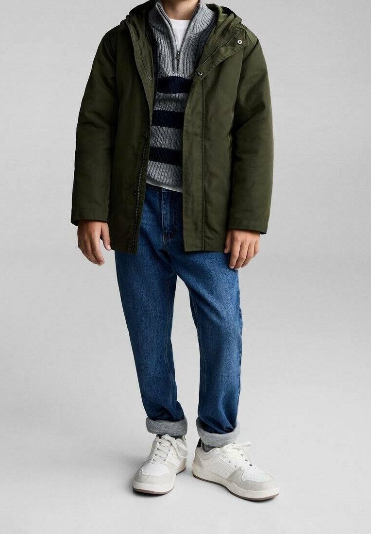 Enfant portant une veste à capuche verte sur un pull rayé gris et bleu marine, jean bleu retroussé et baskets blanches sur fond gris.