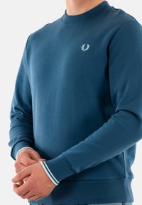 Felpa blu con scollatura a girocollo. Presenta un piccolo logo ricamato sul petto e polsini a righe. Realizzata in tessuto morbido con una superficie liscia.