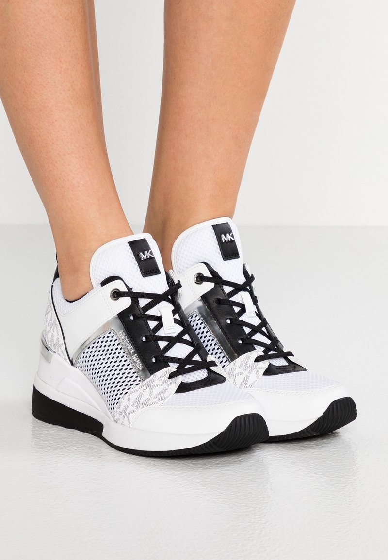 Baskets blanches et noires Michael Kors avec des détails en mesh et motifs, portées pieds nus sur un fond clair uni.