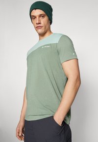 T-shirt verde a maniche corte con pannello spalla verde chiaro, tessuto leggero e logo sul petto; abbinato a pantaloni scuri e un berretto verde.