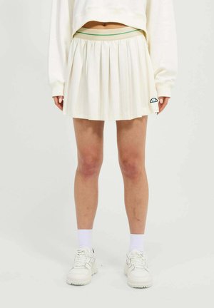 Femme portant une jupe plissée blanche avec un petit logo, des chaussettes blanches, des baskets blanches épaisses et un sweat-shirt court de couleur écrue.