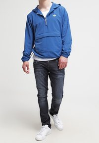 Modrá pulovrová bunda se zipem vpředu, elastickými rukávy a klokáním kapsou. Kombinovaná s tmavými džínami a bílými teniskami. Jednoduchý design.