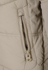 Gai bēšs pūkveida jakse, kurai ir redzama rāvējslēdzēja kabata ar baltām zobiem un bēša krāsas vilkšanas cilpu. Tekstūrēts materiāls ar šuvju detaļām.