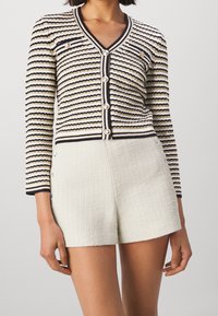 Kobieta w dopasowanej kardiganowej bluzie w paski z dekoltem w serek i złotymi guzikami, w wysokich, fakturowanych białych szortach, stojąca na jednolitym tle.