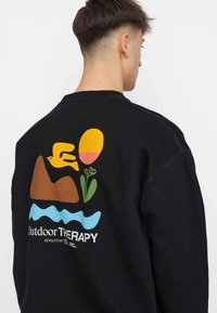 Zwarte sweatshirt met een kleurrijke print op de achterkant van een vogel, berg, bloem, zon en golvend water, met het label "Outdoor Therapy."