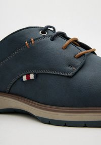 Zapato de cuero azul oscuro con suela beige, cordones marrones y acentos cosidos. Presenta una pequeña etiqueta roja, blanca y azul en el costado.