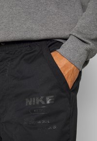 Pantalones negros de Nike con logo reflectante, cinturilla elástica y bolsillos laterales. Puños de sudadera gris visibles por encima de la cinturilla.