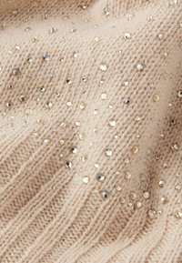 Beżowa dzianina o prążkowanej fakturze, ozdobiona cekinami w różnych rozmiarach, w odcieniach złota i srebra, rozrzuconymi na powierzchni.