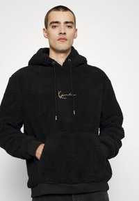 Svart sherpa-hettegenser med en luksuriøs tekstur, justerbare snor med gullender, fremre kengurulomme og brodert logo i gull.