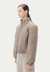 Ljusbrun vadderad jacka med hög krage, dragkedja fram och elastiska ärmslut. Den har en strukturerad form och mjuk tygtextur.