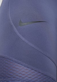 Zbliżenie purpurowej tkaniny sportowej Nike, ukazujące detale przeszyć oraz czarny logo Nike swoosh z siateczkowym panelem na dole.
