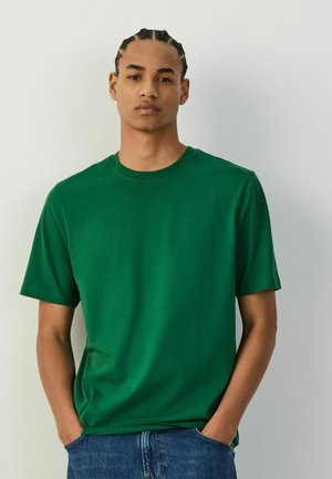 Młody mężczyzna z warkoczami, ubrany w jednolity zielony t-shirt z krótkim rękawem i niebieskie dżinsy, z rękami w kieszeniach, stojący na białym tle.