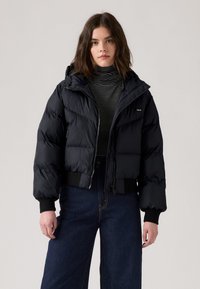 Casaco puffer preto com capuz destacável, design acolchoado, fecho de zip, e punhos canelados, combinado com um suéter de gola alta às riscas e jeans de perna larga.