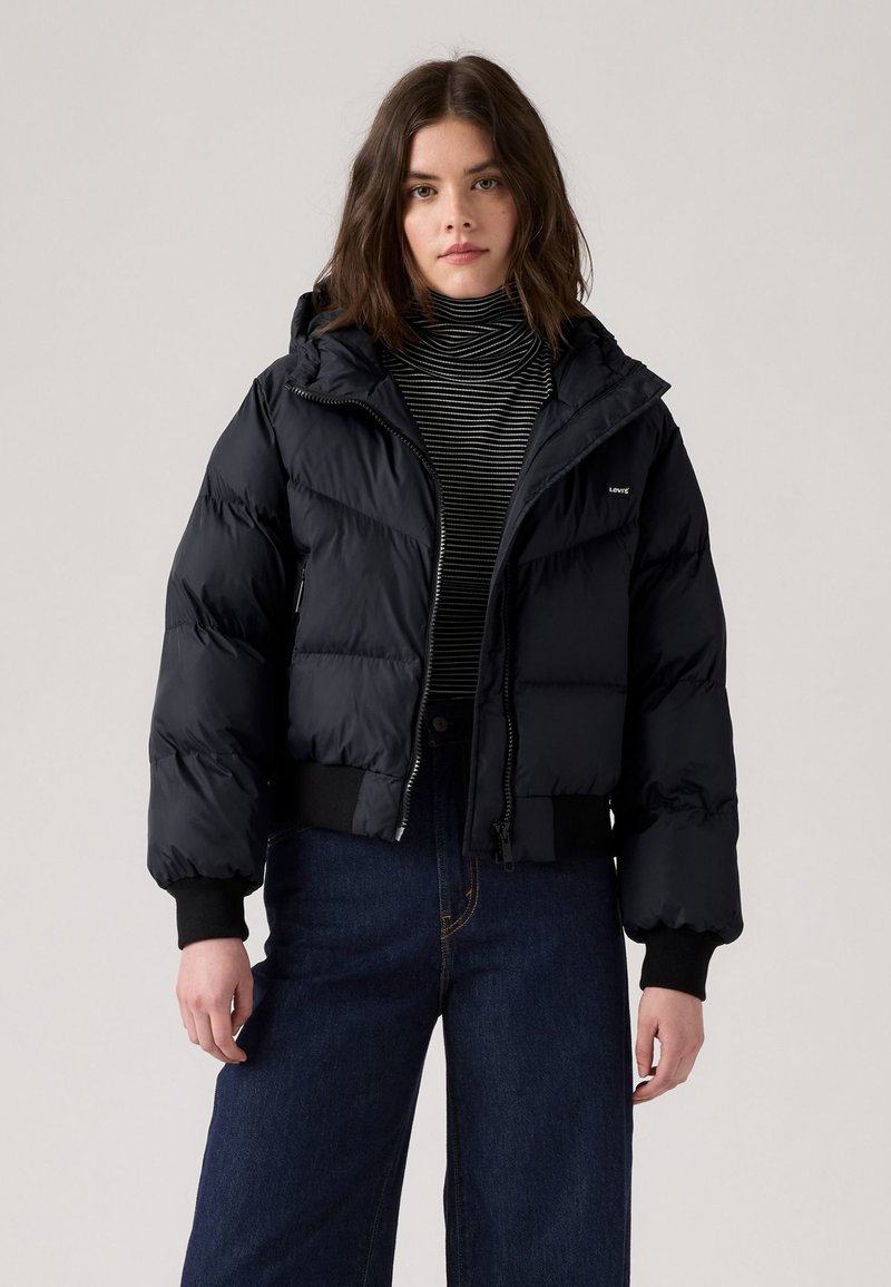 Veste matelassée noire avec capuche amovible, design matelassé, fermeture éclair sur le devant et poignets côtelés, associé à un col roulé rayé et un jean évolué.