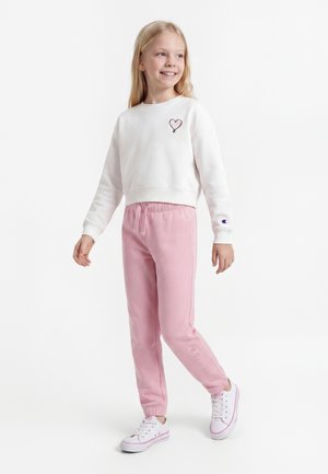 Jeune fille souriante, portant un sweat-shirt blanc avec un patch en forme de cœur, un pantalon de jogging rose avec cordon et des baskets blanches, debout sur fond blanc.