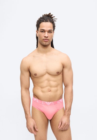 Αθλητικός άνδρας με πλεξούδες που φοράει ροζ εσώρουχα με λευκή ελαστική μέση, στέκεται μπροστά σε απλό ανοιχτό φόντο.