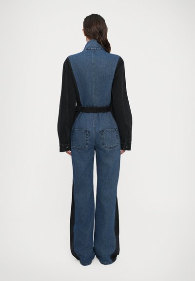 Donna con lunghi capelli scuri che indossa una tuta in denim bicolore con maniche nere e pannelli laterali, in piedi di spalle su uno sfondo bianco.