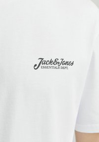 Camiseta de algodón blanca con un logo negro en la parte posterior que dice "Jack & Jones ESSENTIALS DEPT." Diseño de mangas cortas y cuello redondo.