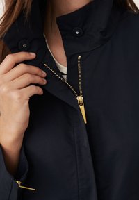 Marineblå jakke med høj krave, guldlukning og knapdetaljer. Stoffet ser glat og let ud med et minimalistisk design.