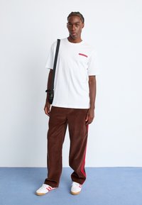 Oversized white cotton t-shirt with a red rectangular logo, paired with brown corduroy pants featuring red side stripes and white sneakers.